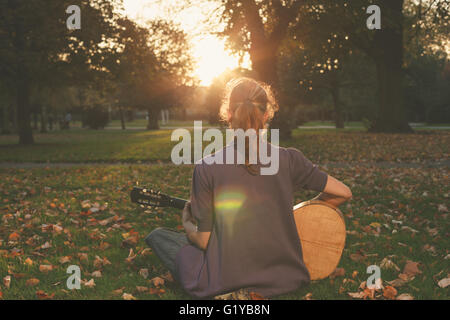 Vue arrière du jeune femme assise sur l'herbe et jouer de la guitare dans le parc au coucher du soleil Banque D'Images