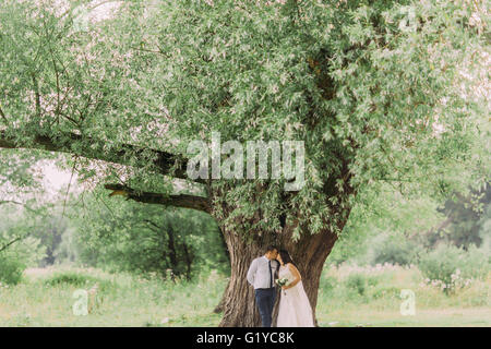 Heureux les jeunes mariés s'embrasser sous un grand arbre avec des feuilles vertes sur spring meadow Banque D'Images