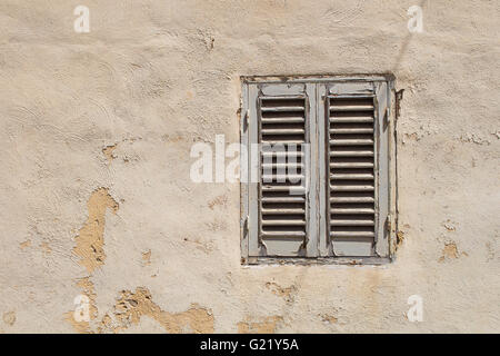 Vieux Mur avec une façade détachée et une petite fenêtre avec un volet fermé. L'ombre d'un câble traversant la fenêtre. Banque D'Images