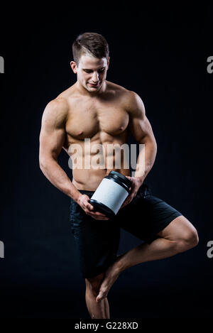 Thème : sport et musculation beau bodybuilder solide en plastique holding pot avec une protéine sèche détendre isolé sur fond noir en studio Banque D'Images