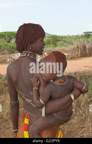 Jeune femme Hamar avec argile rouge dans ses cheveux tenant un bébé dans ses bras et avec les cicatrices et les marques d'être fouetté sur le dos Banque D'Images