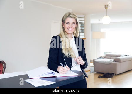 Femme d'affaires d'âge moyen écrit avec un stylo dans une main et tenant une paire de lunettes avec l'autre sourire pour la caméra. Banque D'Images