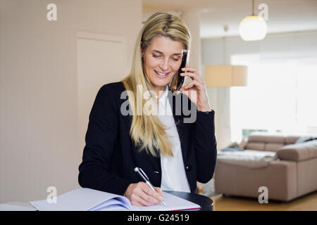 Businesswoman talking on a cell phone et l'écriture avec un stylo. home office. décor propre contemporain. Banque D'Images