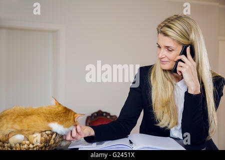 Belle femme d'affaires d'âge moyen à l'aide d'un téléphone et de prendre des notes. Red haired cat sitting in panier tressé à côté d'elle. Se détendre Banque D'Images