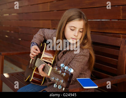 Kid blonde girl learning jouer de la guitare avec béret hiver smartphone sur fond de bois Banque D'Images