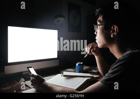 Pensive asian young man studying avec écran blanc ordinateur et smartphone dans la soirée Banque D'Images