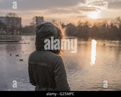 Une personne portant un épais manteau chaud est debout près d'un étang dans un parc et est à regarder le coucher du soleil en hiver Banque D'Images