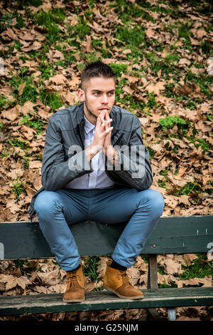 Beau jeune homme assis sur un banc dans le parc en automne, avec les feuilles mortes tout autour de Banque D'Images