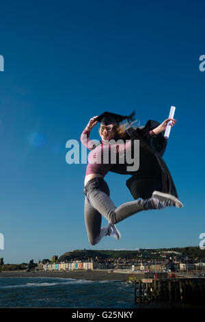 Sautant de joie : un jeune femme fille portant un chapeau d'étudiant de l'université et conseil de mortier sur robe son diplôme date tenant son diplôme sauter de joie dans l'air pour célébrer son succès, avec la ville d'Aberystwyth, Pays de Galles UK dans l'arrière-plan Banque D'Images