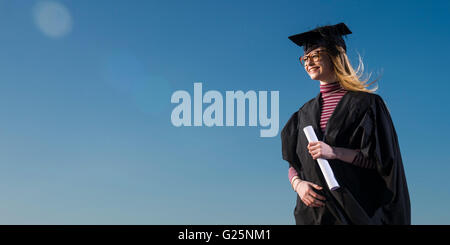 Ciel bleu la réflexion : une jeune femme portant un chapeau d'étudiant de l'université du mortier et une blouse sur son diplôme date tenant son diplôme en contemplant la réflexion sur sa vie future et de carrière après l'enseignement supérieur de l'éducation, UK Banque D'Images