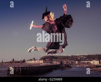 Sautant de joie : un jeune femme fille portant un chapeau d'étudiant de l'université et conseil de mortier sur robe son diplôme date tenant son diplôme sauter de joie dans l'air pour célébrer son succès, avec la ville d'Aberystwyth, Pays de Galles UK dans l'arrière-plan Banque D'Images