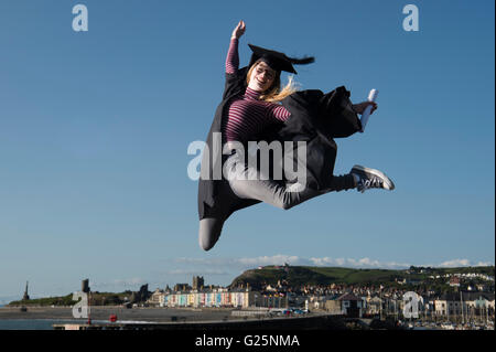 Sautant de joie : un jeune femme fille portant un chapeau d'étudiant de l'université et conseil de mortier sur robe son diplôme date tenant son diplôme sauter de joie dans l'air pour célébrer son succès, avec la ville d'Aberystwyth, Pays de Galles UK dans l'arrière-plan Banque D'Images