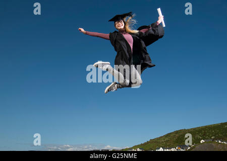 Sautant de joie : un jeune femme fille portant un chapeau d'étudiant de l'université et conseil de mortier sur robe son diplôme date tenant son diplôme sauter de joie dans l'air pour célébrer son succès, UK Banque D'Images