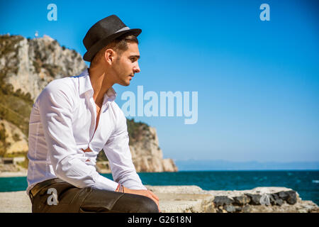 Jeune homme séduisant en costume classique sur beach holding lunettes de soleil pour détourner la tête. Les vagues de la mer sur l'arrière-plan Banque D'Images