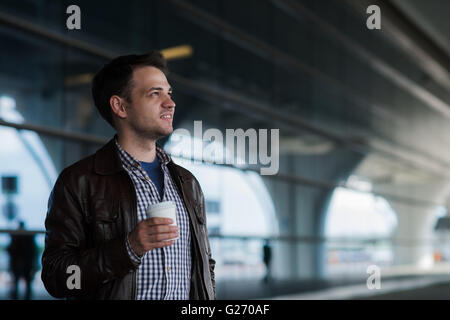 Beau jeune homme élégant en soie de voyageur avec l'extérieur permanent près de l'aéroport. Homme portant veste et chemise. Personne souriant à la caméra à holding tasse de café Banque D'Images