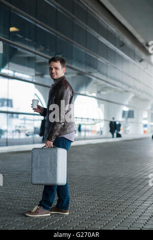 Portrait de profil d'un jeune homme marchant avec valise et tasse de café près de l'aéroport Banque D'Images