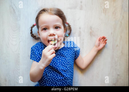 Petite fille allongée sur le sol, smiling, manger une sucette Banque D'Images