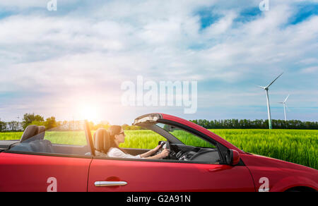 Girl in red cabriolet dans un champ avec des éoliennes au coucher du soleil. Banque D'Images