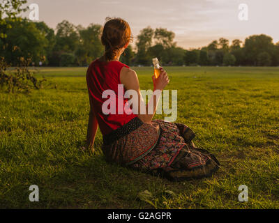 Une jeune femme est en train de boire d'une bouteille en position assise sur l'herbe dans un parc et d'admirer le coucher du soleil Banque D'Images