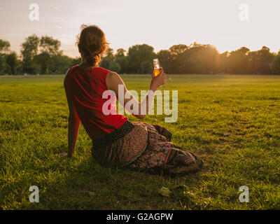 Une jeune femme est en train de boire d'une bouteille en position assise sur l'herbe dans un parc et d'admirer le coucher du soleil Banque D'Images