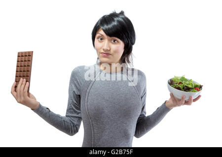 Jeune, jolie modèle fitness essaie de décider entre une grande barre de chocolat et un bol de salade verte. Banque D'Images