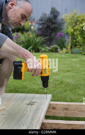 Un ouvrier utilise une perceuse sans fil pour assembler de terrasse de jardin dans un jardin doté d''une pelouse et d'arbustes dans l'arrière-plan Banque D'Images