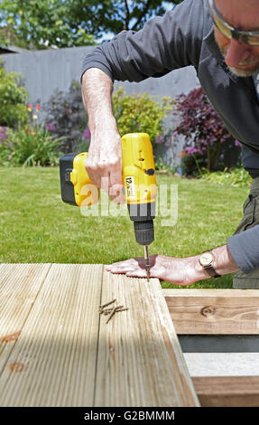 Un ouvrier utilise une perceuse sans fil pour assembler de terrasse de jardin dans un jardin doté d''une pelouse et d'arbustes dans l'arrière-plan Banque D'Images