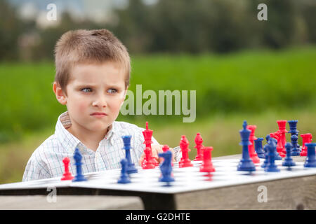 Jeune garçon élégant en chemise blanche d'apprendre à jouer aux échecs avec des pièces bleu et rouge sur la table en bois dans le parc Banque D'Images