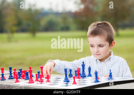 Jeune garçon élégant en chemise blanche d'apprendre à jouer aux échecs avec des pièces bleu et rouge sur la table en bois dans le parc Banque D'Images