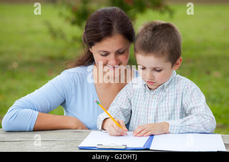 Peu de jeune garçon en chemise blanche avec sa mère enseignante, écrire ou dessiner avec un crayon sur une feuille de papier sur bois tableau i Banque D'Images