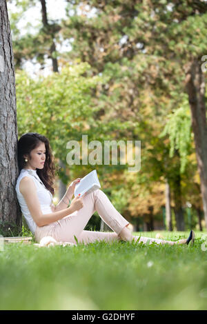 Les jeunes d'Asie femme appuyée contre un arbre et assis dans l'herbe lire un livre dans le parc Banque D'Images