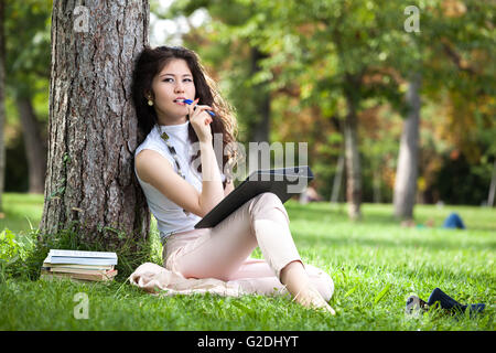 Jeune femme stand asiatique appuyé contre un arbre dans le parc et de penser ce qu'il faut écrire dans son programme Banque D'Images