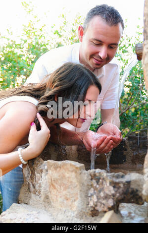 Jeune couple habillé en blanc en face d'une fontaine où le marié donne à l'épouse de boire avec les mains.Vertical Banque D'Images