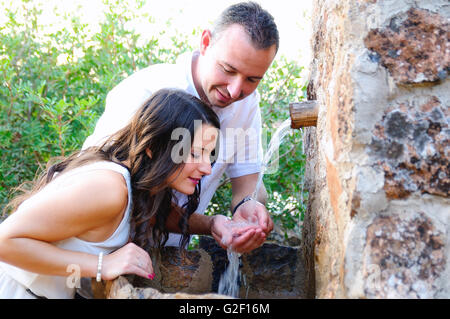 Jeune couple habillé en blanc en face d'une fontaine où le marié donne à l'épouse de boire avec les mains Banque D'Images