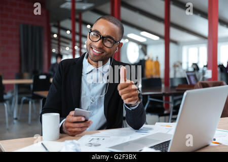 Jeune homme élégant montrant bien signer et holding smartphone à l'ordinateur portable Banque D'Images