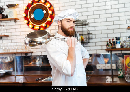 Chef cuisine attrayante grave debout et tenant poêle sur la cuisine Banque D'Images