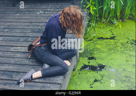 Une jeune femme est assise sur une terrasse en bois par un étang sale avec des algues dans une forêt Banque D'Images