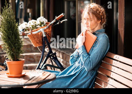 Thoughtful young woman with bicycle livre readng et rêver sur le plein air beanch Banque D'Images
