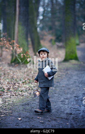 Mignon petit enfant, garçon d'âge préscolaire, tenant une lanterne et de livre, marcher dans une forêt sombre Banque D'Images