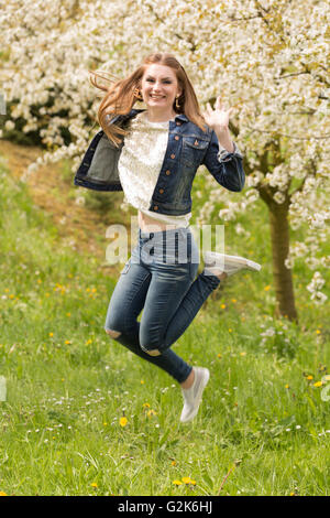 Une jeune femme brune en jeans profitant des premiers jours du printemps dans un pré vert entouré de cerisiers en fleurs Banque D'Images