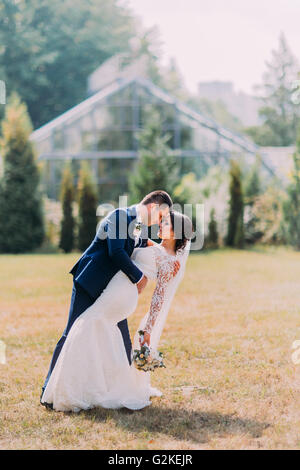 Belle Mariée avec une superbe robe de mariée blanche et le marié en costume bleu élégant posant en plein air, sur la pelouse. L'arrière-plan à effet de serre Banque D'Images