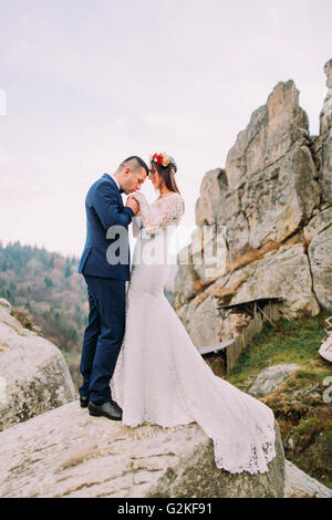 Beau palefrenier en costume bleu élégant embrassant amoureusement ses mains sur mariée blanc habillé de montagnes avec de gros rochers comme contexte Banque D'Images