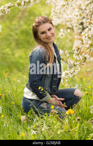 Une jeune femme brune en jeans profitant des premiers jours du printemps dans un pré vert entouré de cerisiers en fleurs Banque D'Images