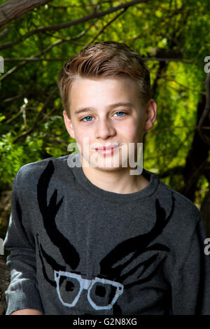 Portrait d'un garçon tween s'appuyant sur un arbre avec une expression arrogant. Il a de gros, les yeux bleus, cheveux blonds, et porte un pull Banque D'Images