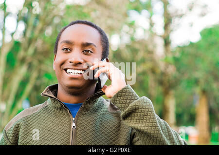 Handsome young black man wearing green chandail en dehors parc salon holding up phone et parler en riant joyeusement Banque D'Images