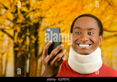 Les jeunes blessés positive black hispanic male wearing neck brace et souriant, tenant dans la prise de téléphone cellulaire comme jaune, selfies abstract background Banque D'Images