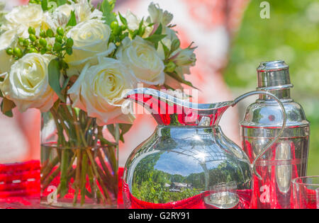 Nature morte à la cruche d'argent et d'argent pour cocktail, fresh cut roses dans vase en verre avec de l'eau sur la table rouge Banque D'Images
