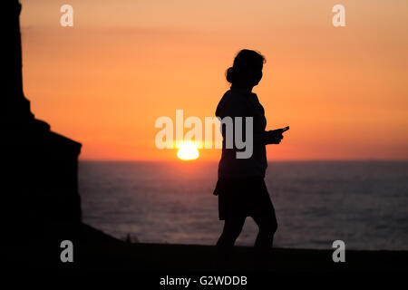 Météo France : une jeune femme marchant avec son téléphone portable dans la main alors que le soleil se couche sur l'horizon sur la côte ouest du pays de Galles. Crédit : Ian Jones/Alamy Live News Banque D'Images