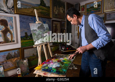 Peintre mâle pensif tient des pinceaux et de la peinture par paletteknife de mélange sur la palette de couleurs de peinture à l'huile mélangée dans un studio avec fond d'œuvres d'accroché au mur. Banque D'Images