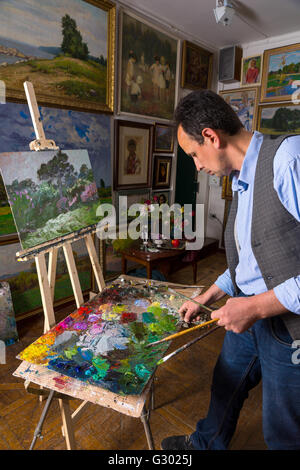 Artiste masculin pensif tient des pinceaux et de la peinture par paletteknife de mélange sur la palette de couleurs de peinture à l'huile mélangée dans une galerie avec des œuvres d'arrière-plan accroché au mur. Banque D'Images